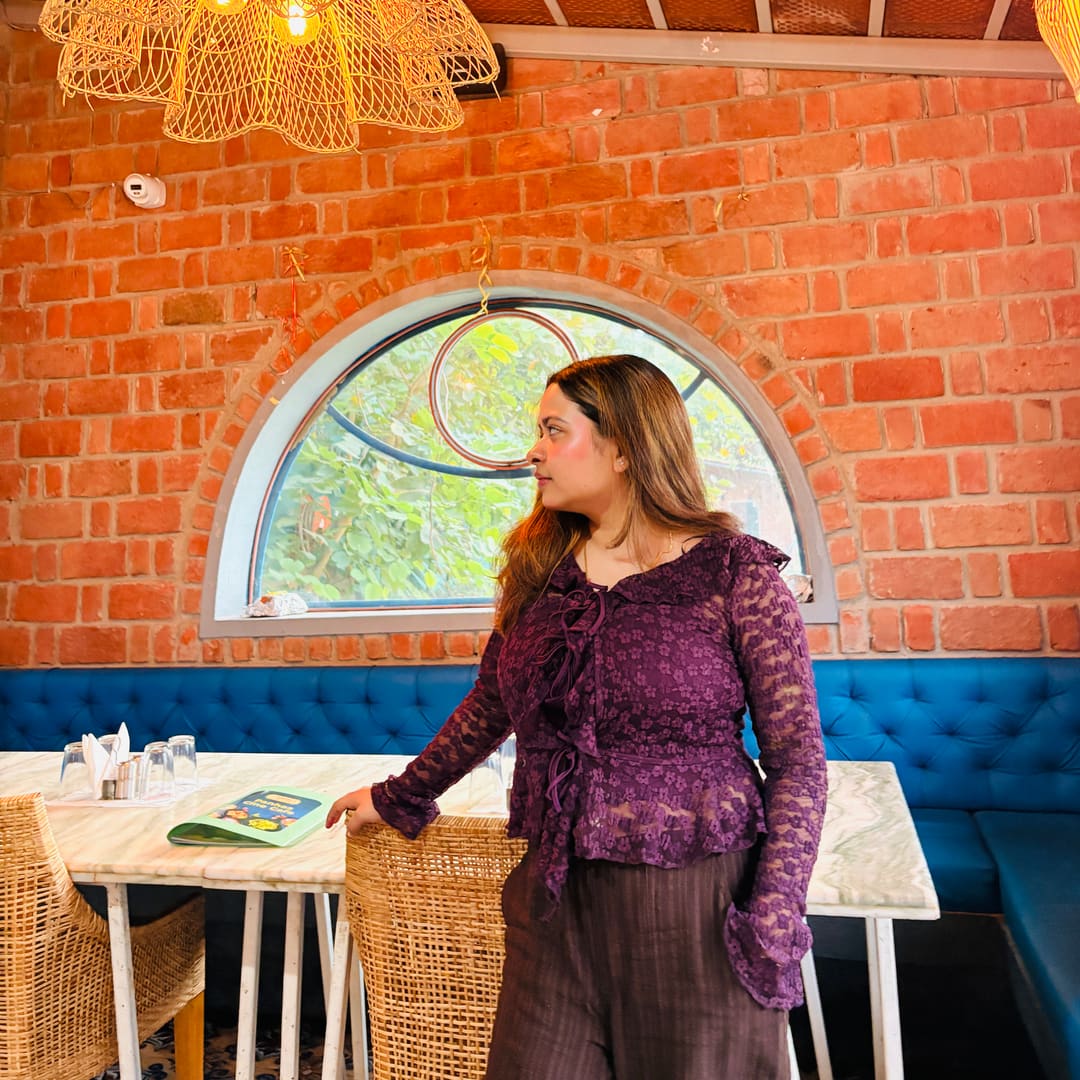 Soumya Singh standing by a woven rattan chair under a hanging cane lamp in a brick café