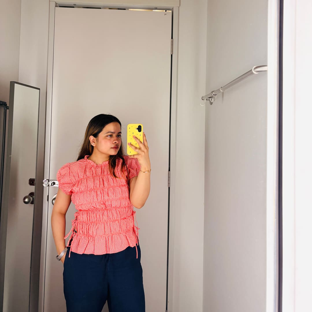 Soumya Singh in a coral gingham top and navy trousers against a pale grey wall
