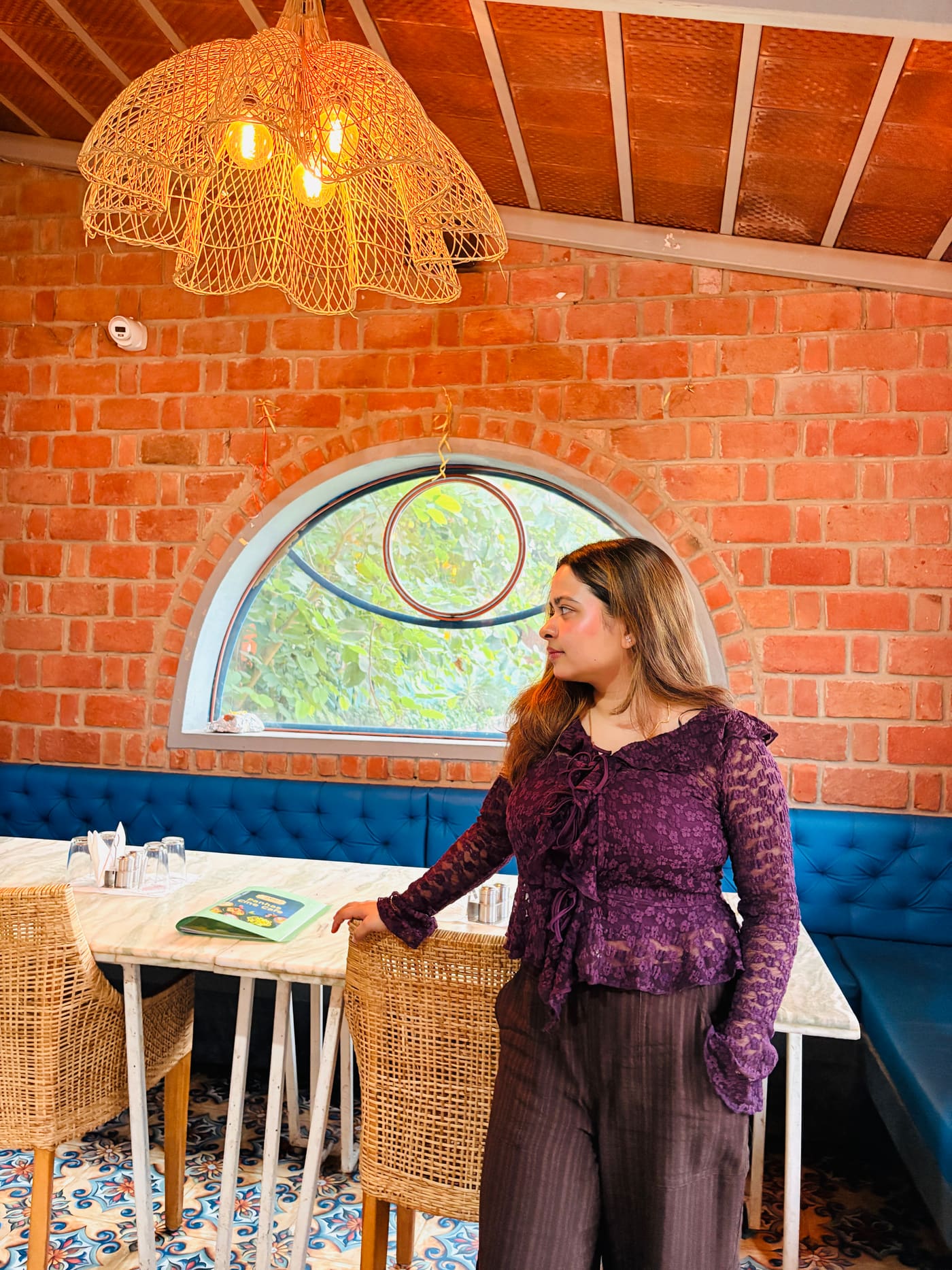 Soumya Singh in a purple lace top, standing in a brick-walled café beside an arched window